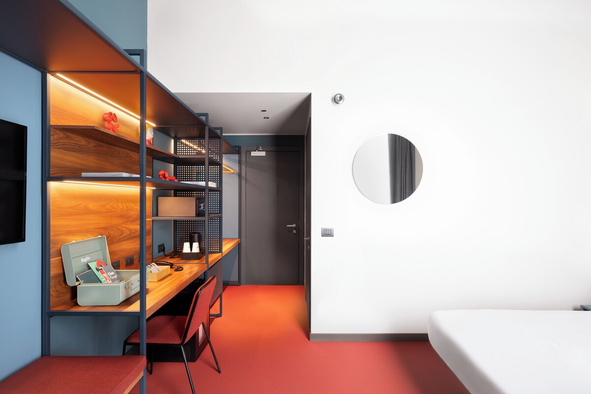 A modern room with a desk, chair, and a bed. There is a shelf unit with various items and a circular mirror on the wall.