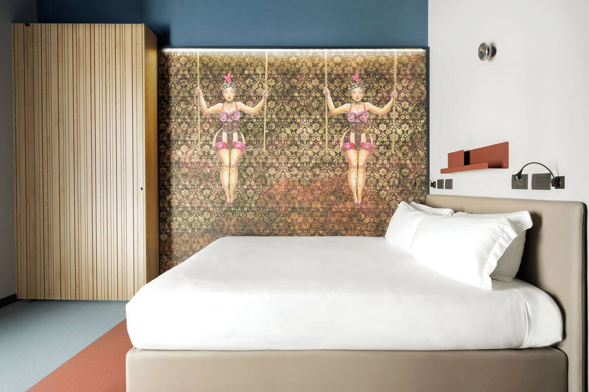 A hotel room with a bed, a wooden panel wall, and a patterned wallpaper with two figures.