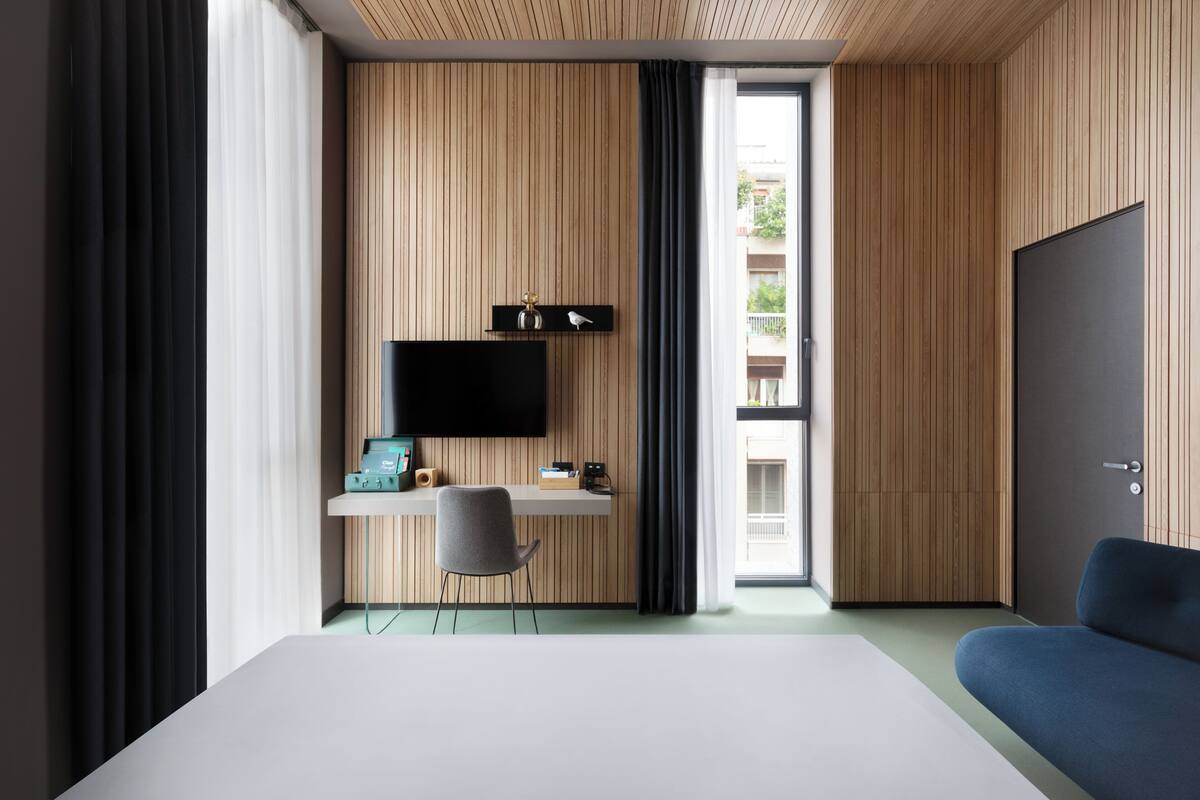A modern hotel room with a bed, a desk with a flat-screen TV, a chair, and a view of the outside.