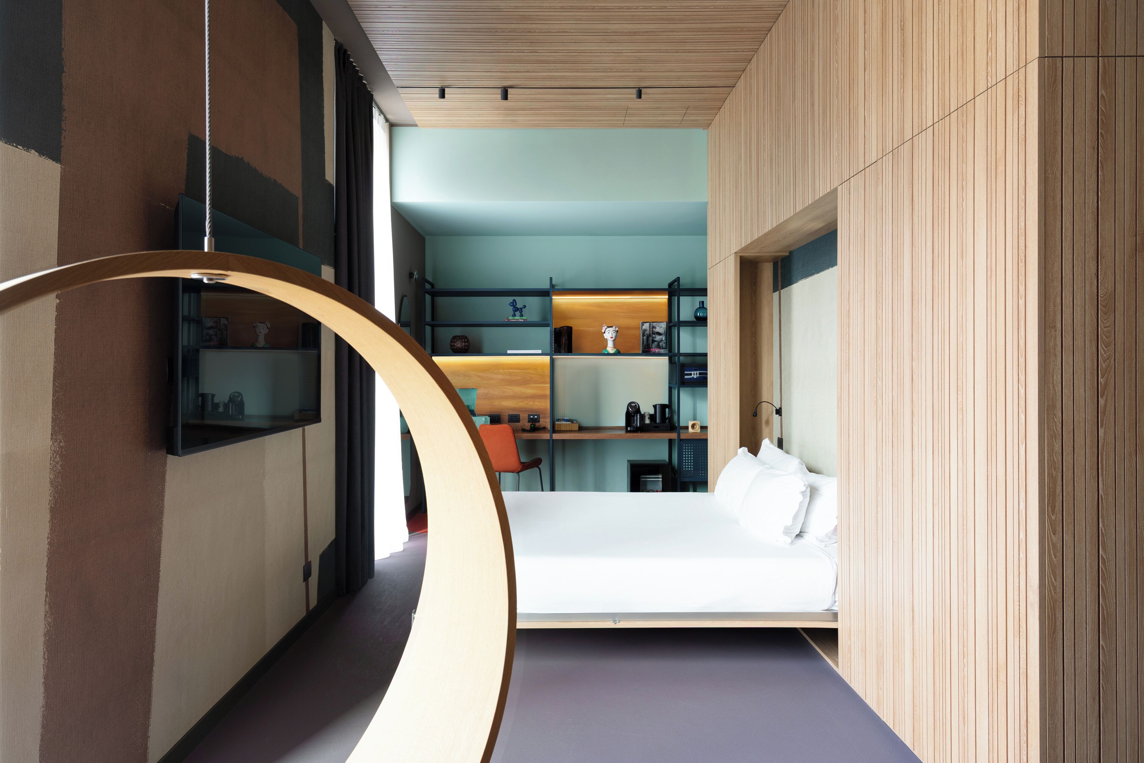 Une chambre d’hôtel moderne dotée d’une structure en bois aux lignes courbes, d’un lit, d’un bureau et d’une étagère contenant divers objets.