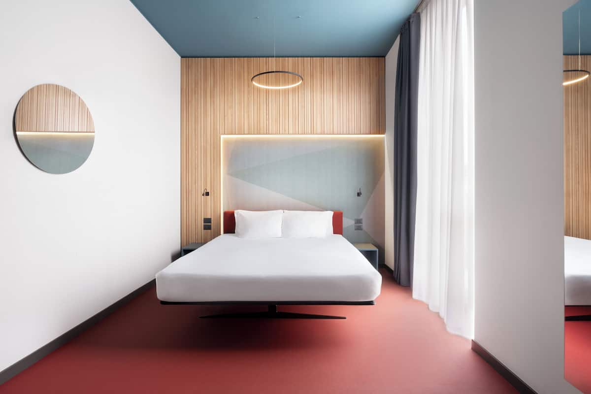 A modern hotel room with a bed, a round mirror, a headboard with built-in lighting, and a red carpet.