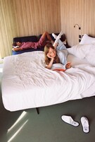 Een bed met wit beddengoed, iemand ligt erop en een paar slippers op de vloer.