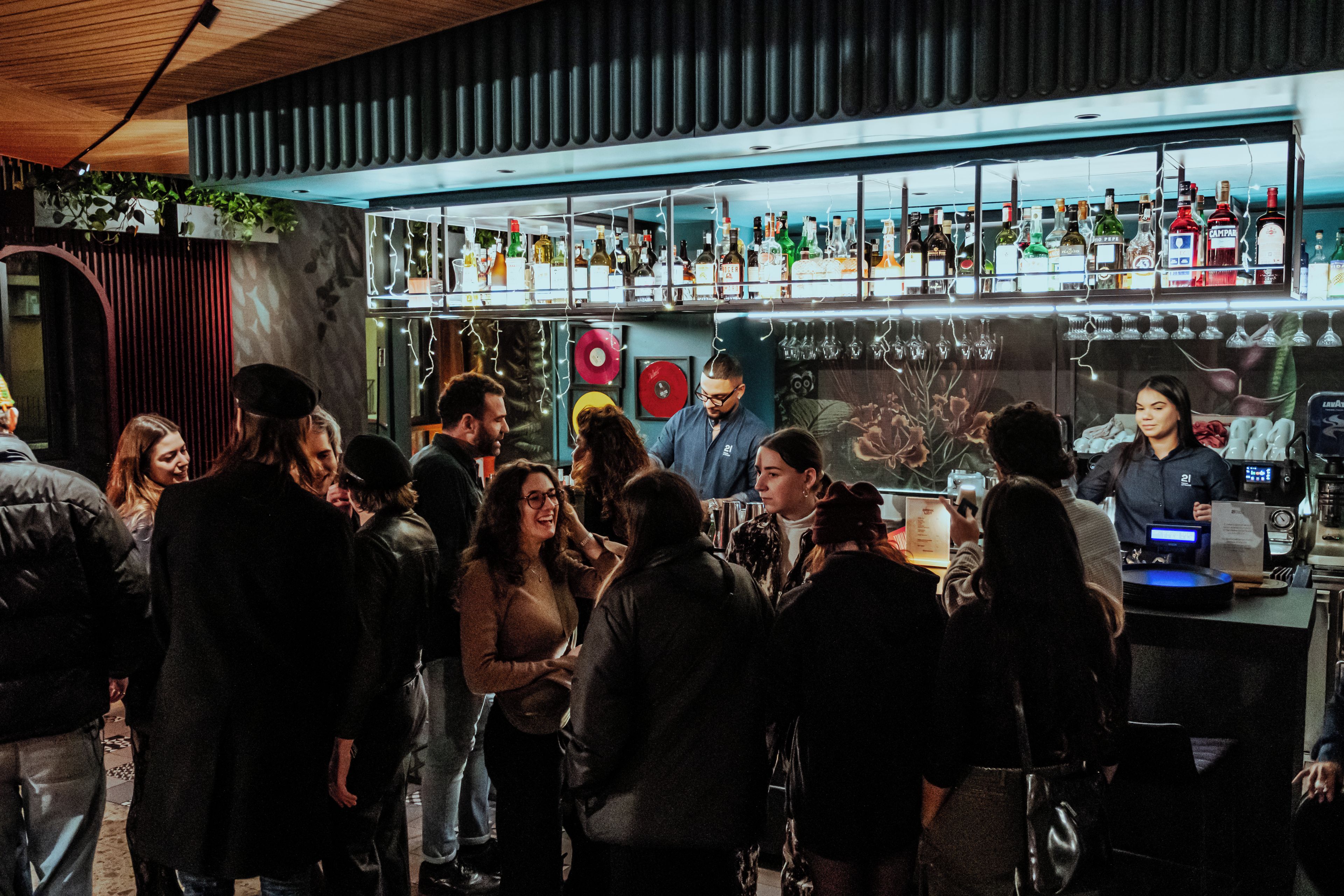 A bar area with a crowd of people, shelves with bottles, and a counter with a person behind it.