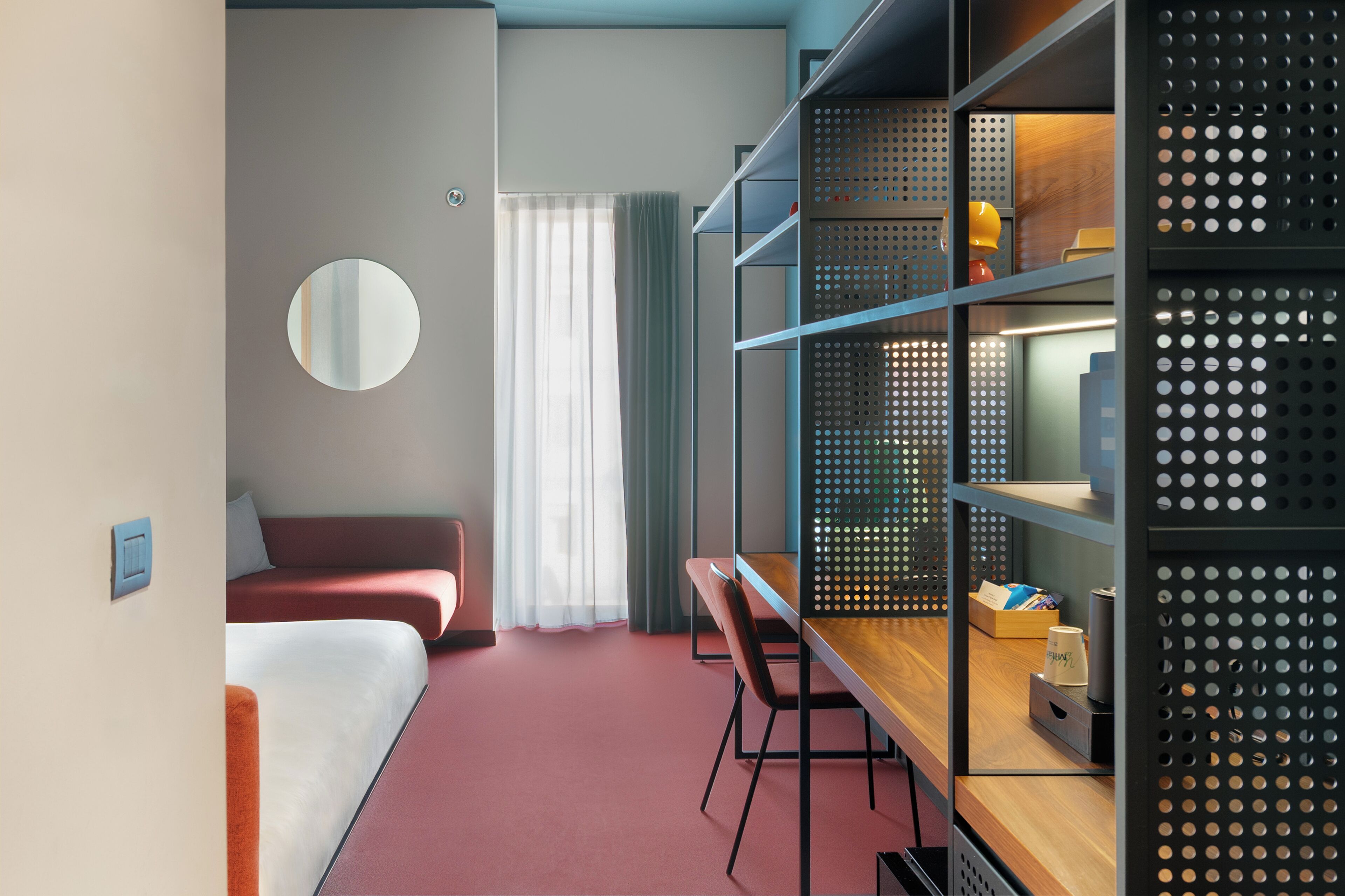 Habitación de hotel con cama, escritorio, silla, espejo redondo y estantería con diseño perforado.