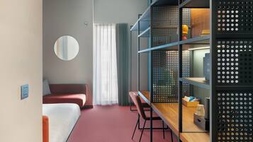 Habitación de hotel con cama, escritorio, silla, espejo redondo y estantería con diseño perforado.