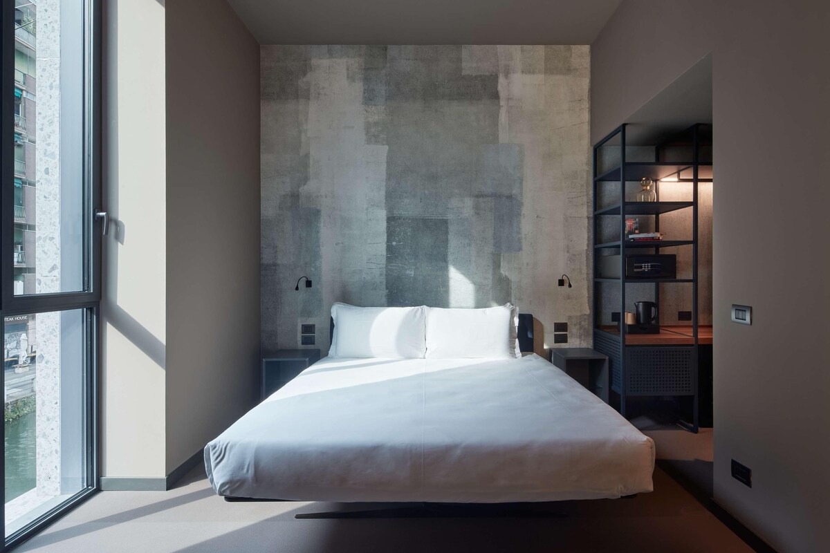 A modern bedroom with a large bed, a concrete accent wall, and a built-in bookshelf.