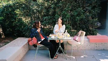 Two people seated at an outdoor table with a meal, surrounded by greenery.