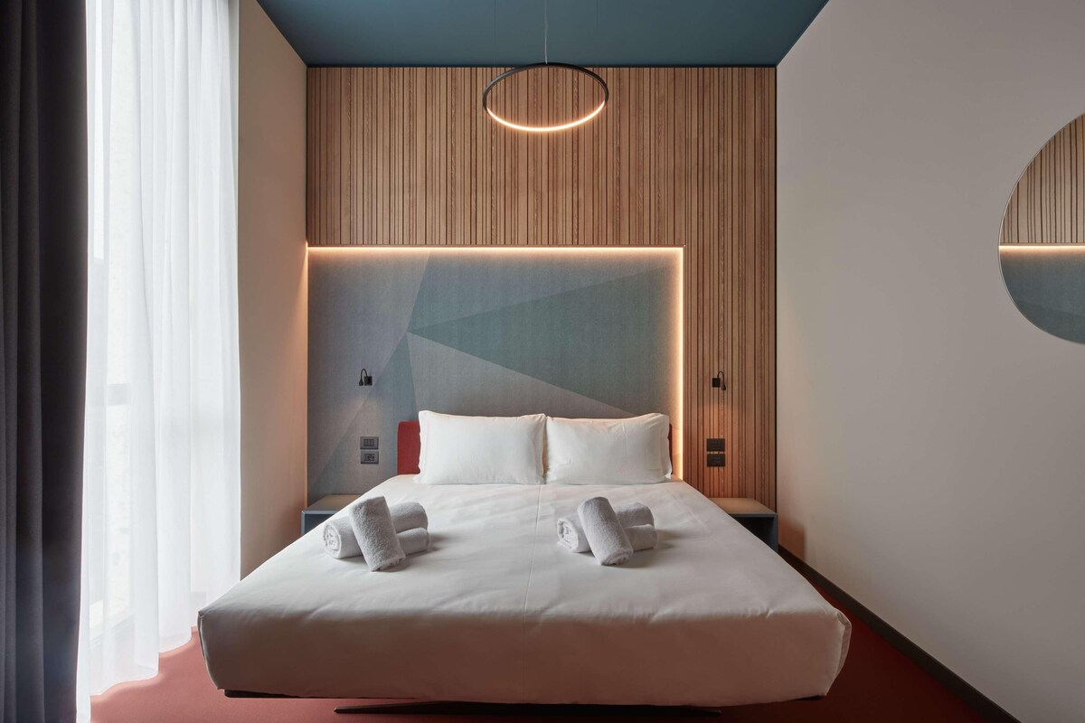 A modern hotel room with a large bed, a wooden panel wall, and a circular light fixture.