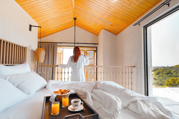 Habitación de hotel con techo de madera, balcón con vistas al campo, cama con ropa blanca y una bandeja con productos para el desayuno.