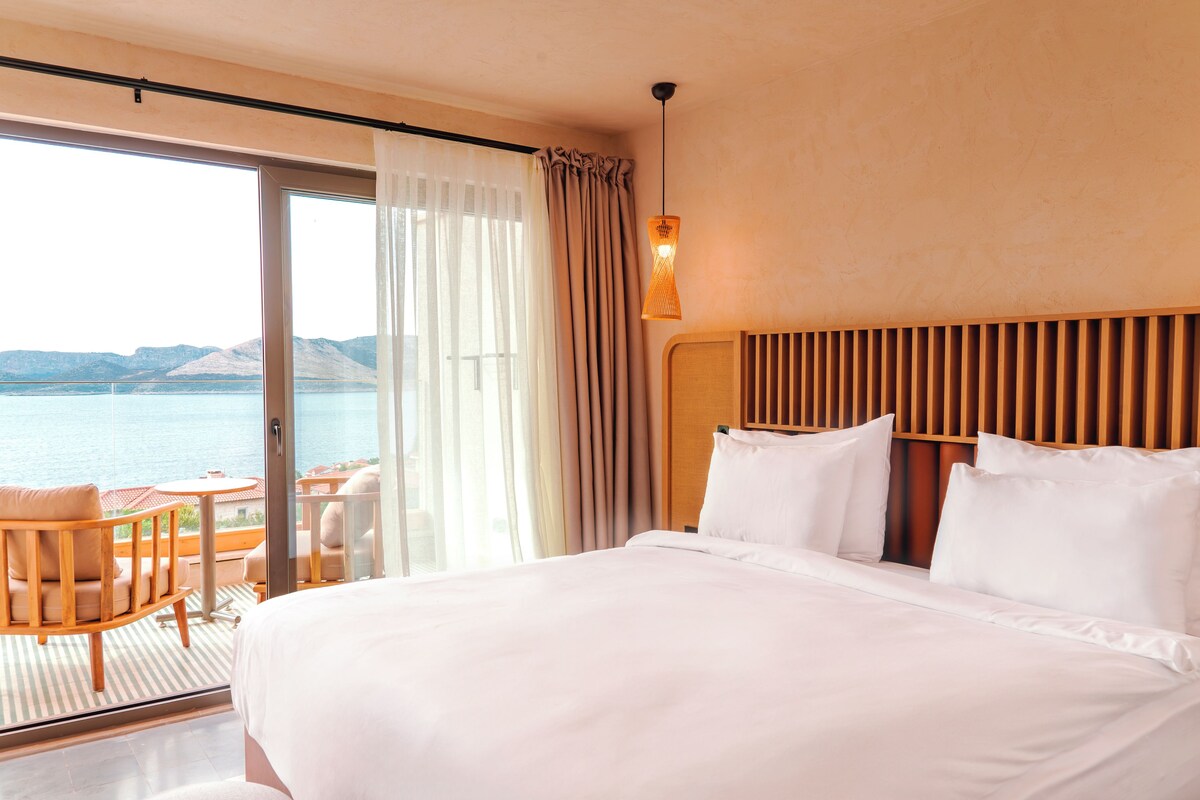 Habitación de hotel con una cama grande, cabecera de madera, balcón con mesa y sillas, y vista al mar.