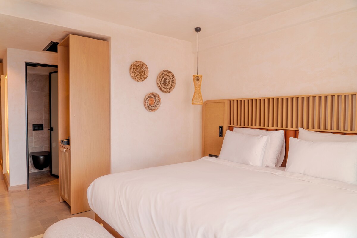 Una habitación de hotel moderna con una cama grande, cabecera de madera y un baño pequeño visible a través de una puerta abierta.