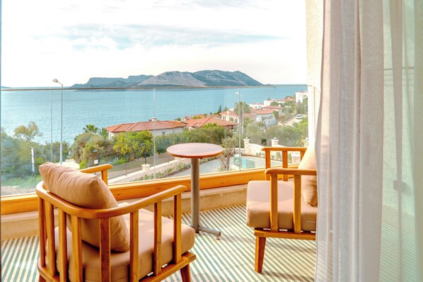 A balcony with wooden chairs and a small table, overlooking a body of water and distant mountains.