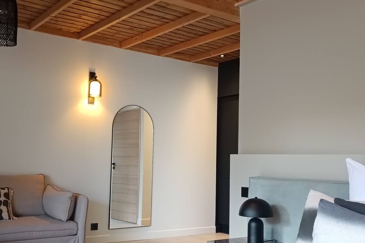 Une chambre avec un plafond en bois, un lit avec un matelas noir, un canapé gris et un luminaire fixé au mur.