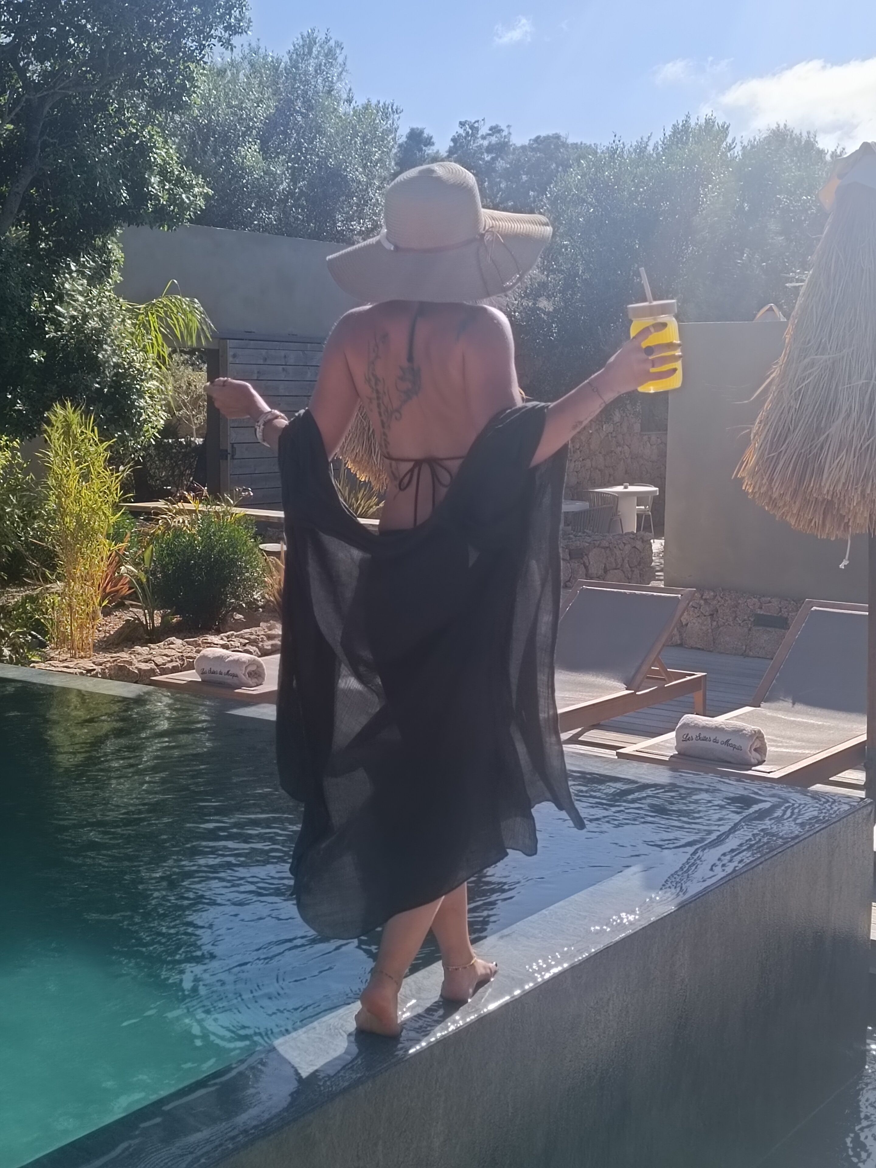 Una persona de pie junto a una piscina, sosteniendo una bebida, usando sombrero y vestido negro, con sillones de playa y un techo de paja al fondo.