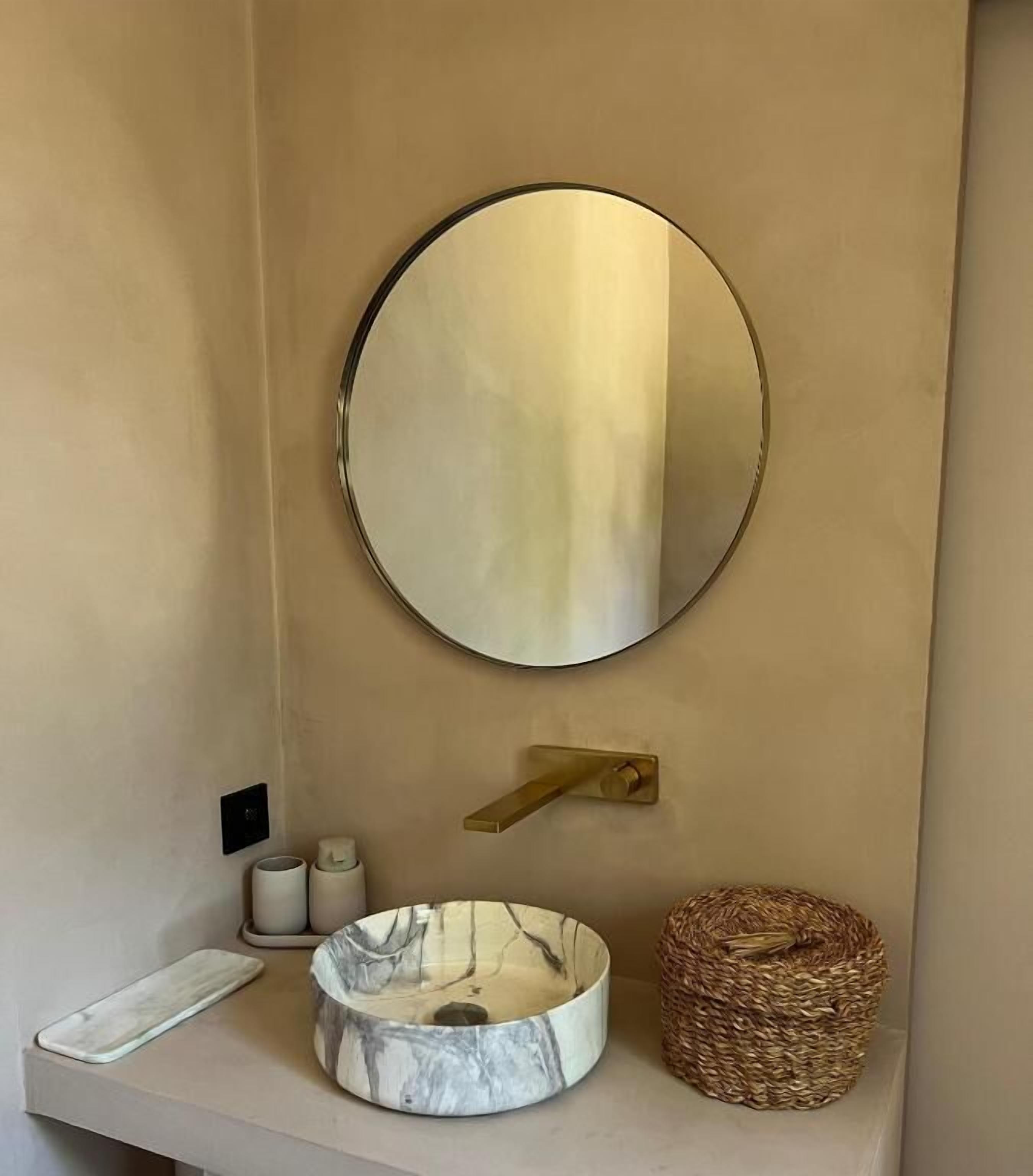 Un bagno con uno specchio rotondo, un lavabo in marmo, un piccolo cestino e un dispenser per asciugamani.