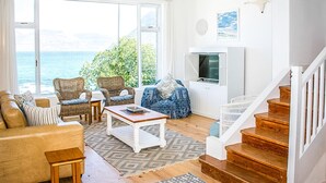 Ein geräumiges Wohnzimmer mit großem Fenster mit Meerblick, einer weißen Treppe und einem Fernseher an der Wand.