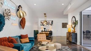 A modern living room with a blue sofa, a brown leather chair, and a round coffee table. There is a dining area in the background with a chandelier. - The Compass Rose: Luxury home, Hot Tub, Pickleball (Hurricane)