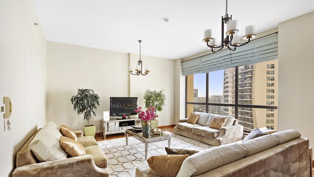 A spacious living room with a large sofa, a coffee table, and a TV. There is a large window with a view of tall buildings. - WelHome - Waterfront Family-Size Apt With JBR Beach Access (Dubai)