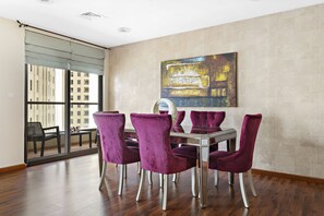 Zona pranzo con tavolo in vetro e sedie viola, un grande quadro astratto sulla parete, e vista su un balcone esterno attraverso una porta in vetro.