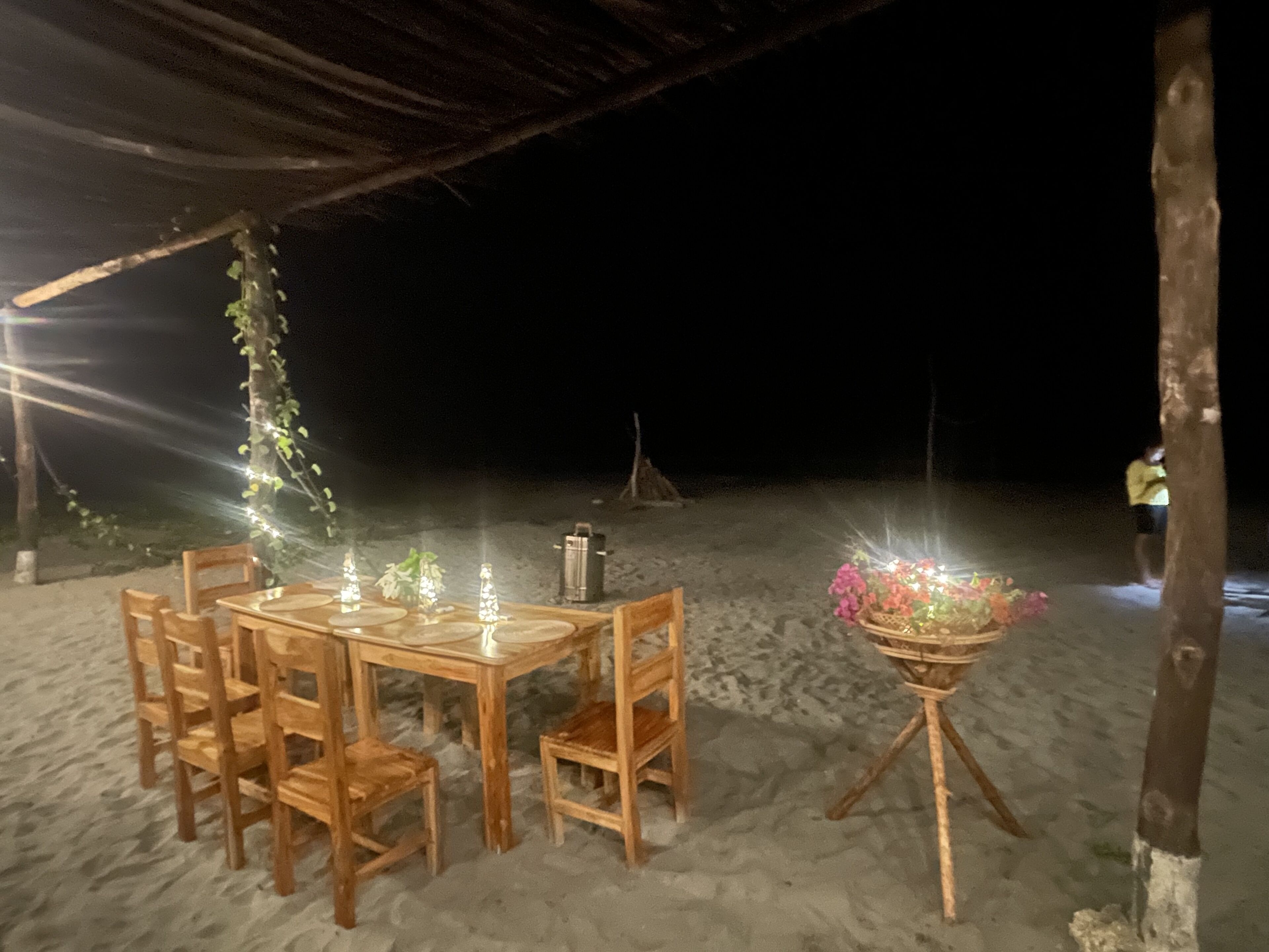A wooden table and chairs set up on a sandy beach at night, with string lights and a floral arrangement.