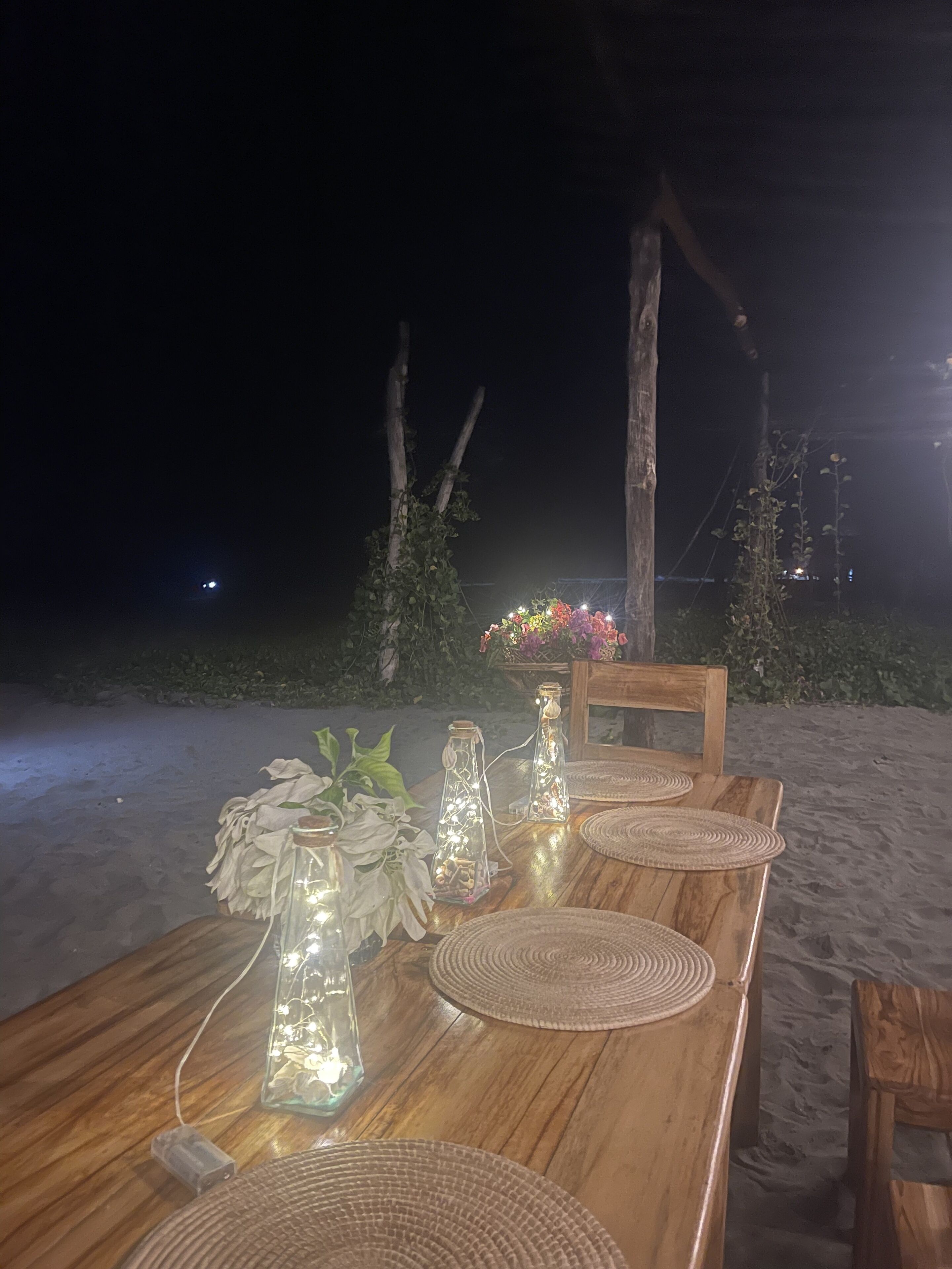 Mesa de madera con individuales tejidos y botellas de vidrio con luces, en el exterior de noche.
