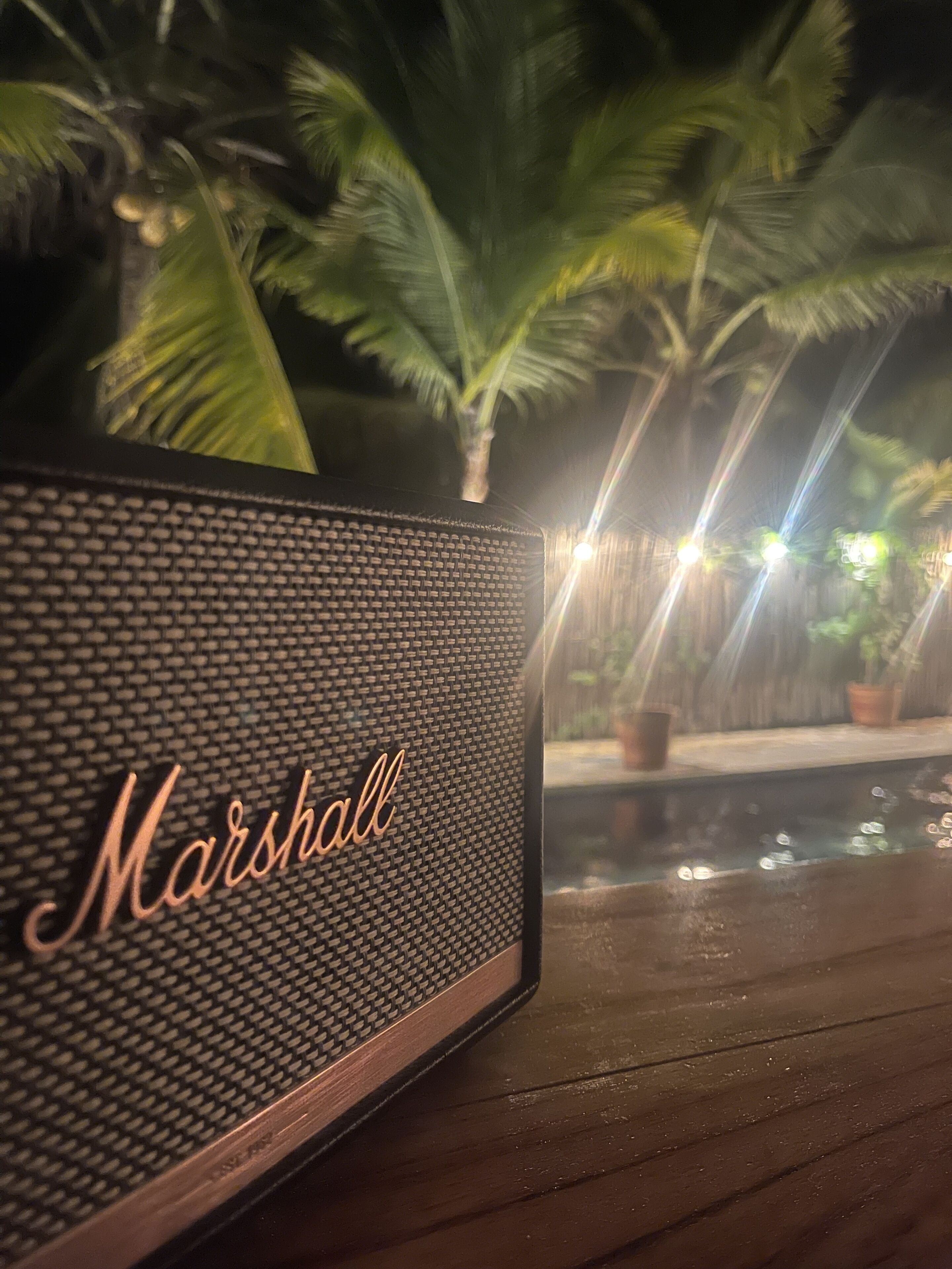 Een houten tafel met een zwarte speaker met het label 'Marshall' en een zwembad met palmbomen op de achtergrond.