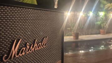 Een houten tafel met een zwarte speaker met het label 'Marshall' en een zwembad met palmbomen op de achtergrond.