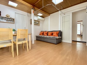 A modern living room with a wooden dining table, two wooden chairs, a grey sofa with orange cushions, and a ceiling with skylights.