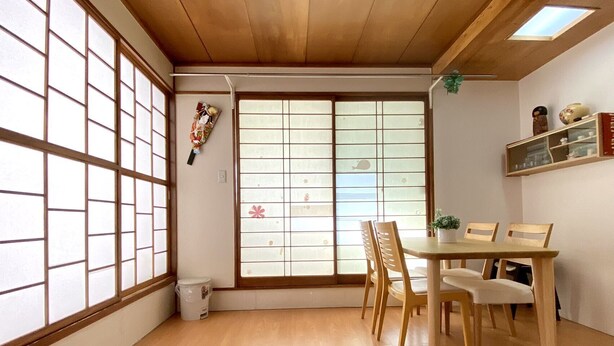 Una habitación japonesa tradicional con suelo de tatami, una puerta corredera shoji, una mesa de madera con sillas y un pequeño estante con objetos decorativos.