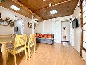 A dining area with a wooden table and chairs, a grey sofa with orange cushions, and a television mounted on the wall.