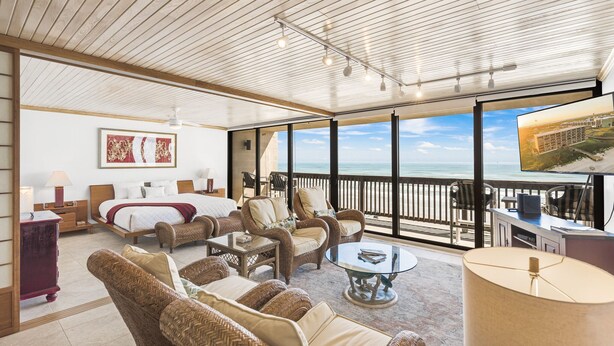 A spacious room with a large bed, two armchairs, a sofa, and a glass coffee table. There is a television on a stand and a large window with a view of the ocean. - Gulf Shores Condo 509 Beachfront Views & Coastal Gem (Port Aransas)