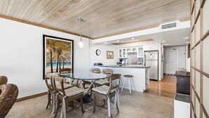 A modern kitchen with white cabinets, a central island, and a dining area with a glass table and wicker chairs. - Gulf Shores Condo 509 Beachfront Views & Coastal Gem (Port Aransas)