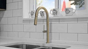 A kitchen with a stainless steel sink, a gold faucet, and a window with white tiles and decorative mugs.