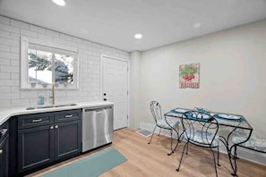 Une cuisine moderne avec des carreaux de céramique blancs disposés en motif métro, un lave-vaisselle en acier inoxydable et un coin repas comprenant une table en verre et des chaises en métal.