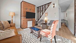 A living room with a brick fireplace, a flat-screen TV, a coffee table, and a staircase leading to another room.