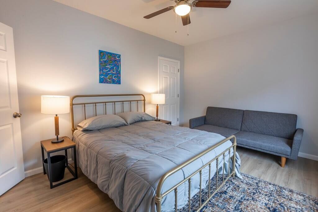 Une chambre à coucher avec un lit, des lampes de chevet, un canapé gris, un ventilateur de plafond et un tableau abstrait bleu accroché au mur.