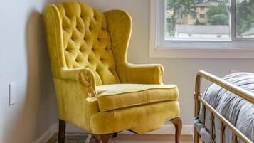 A yellow armchair with a tufted backrest in a room with a window, a bed with a metal frame, and a wall-mounted shelf.