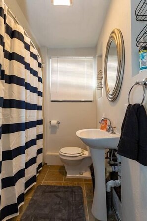 Une salle de bain avec un rideau de douche à rayures, un lavabo blanc, des toilettes et une fenêtre avec des stores.