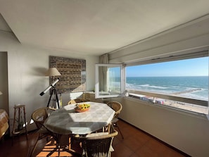 A room with a view of the ocean, a dining table with chairs, a lamp, and a wall decoration.
