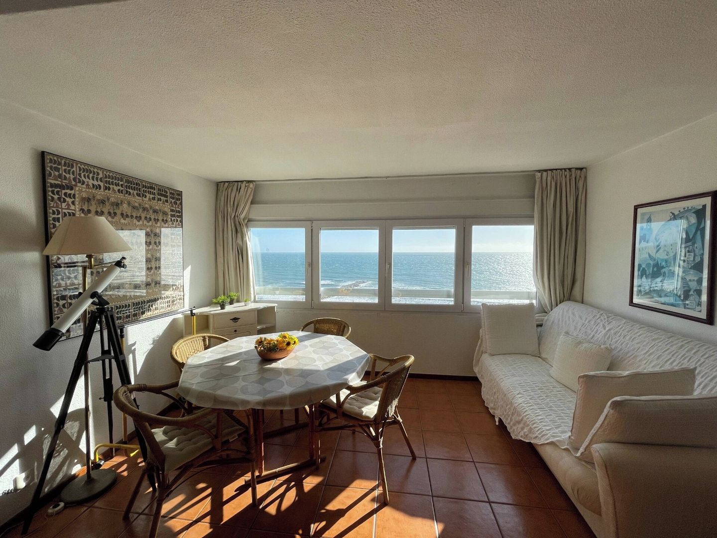 A room with a dining table, chairs, a sofa, a telescope, and a large window with a view of the sea.
