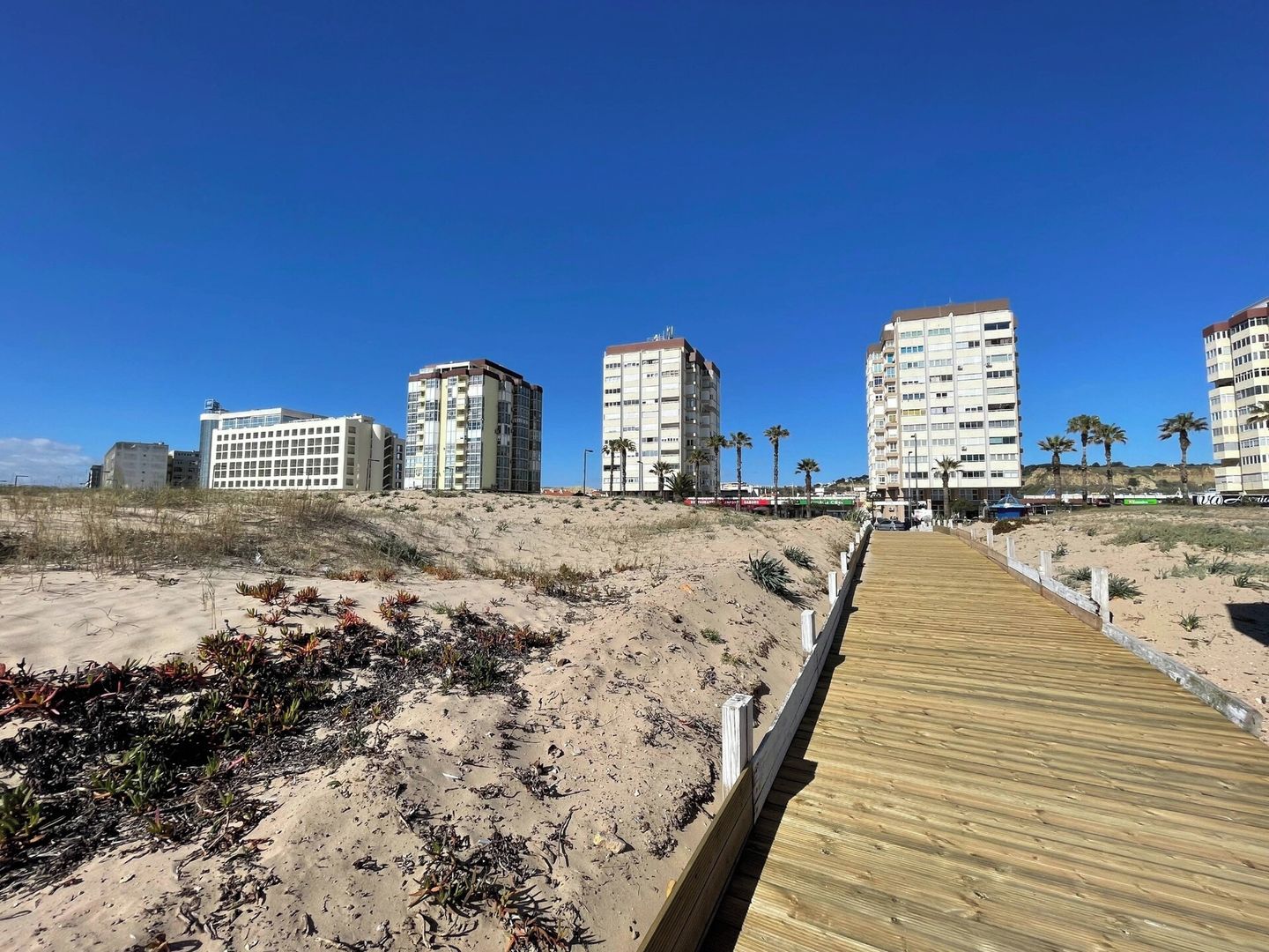 A wooden boardwalk leading to beachfront buildings with a clear sky.