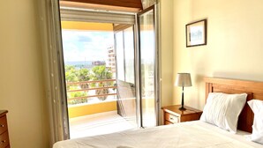 Habitación de hotel con cama, mesita de noche, lámpara, un cuadro en la pared y vistas a un balcón con plantas y edificios.