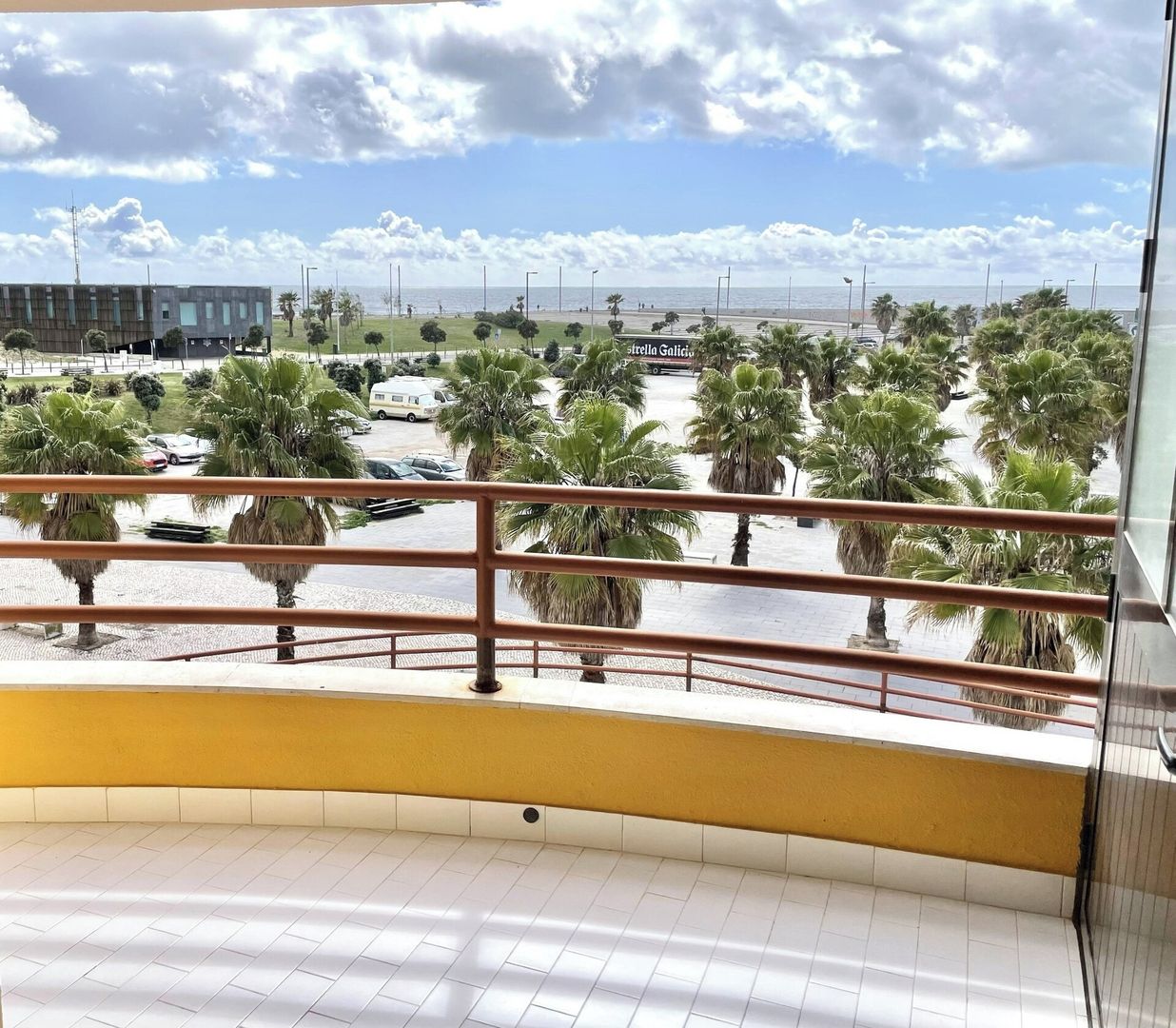 Un balcón con vista a una playa, palmeras y un edificio al fondo.