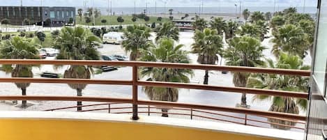 Un balcón con vista a una playa, palmeras y un edificio al fondo.