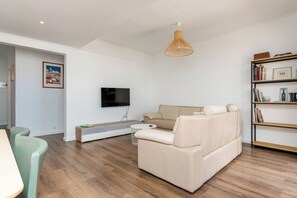 A modern living room with a sofa, a television, a bookshelf, and a dining area with chairs. - Charming 2-bedroom apartment in Caparica (Almada)