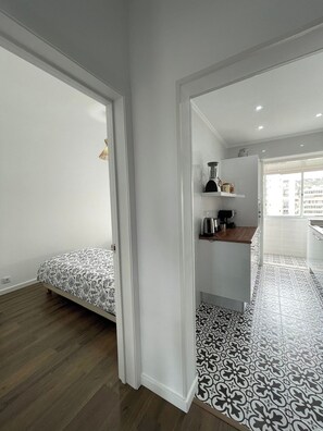 A modern, minimalist room with a bed, a kitchenette, and a view of the city. - Charming 2-bedroom apartment in Caparica (Almada)