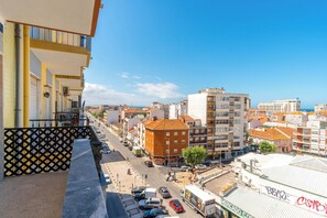 A balcony overlooking a city street with buildings, a market, and parked cars. - Charming 2-bedroom apartment in Caparica (Almada)