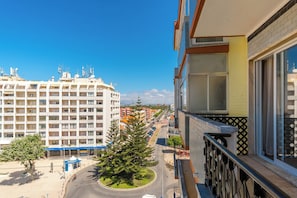 A high-rise building with multiple floors, a balcony with a patterned railing, and a view of a street with parked cars and a tree.