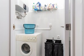 A laundry room with a washing machine, a dryer, a basket, and a shelf with cleaning products. - Charming 2-bedroom apartment in Caparica (Almada)