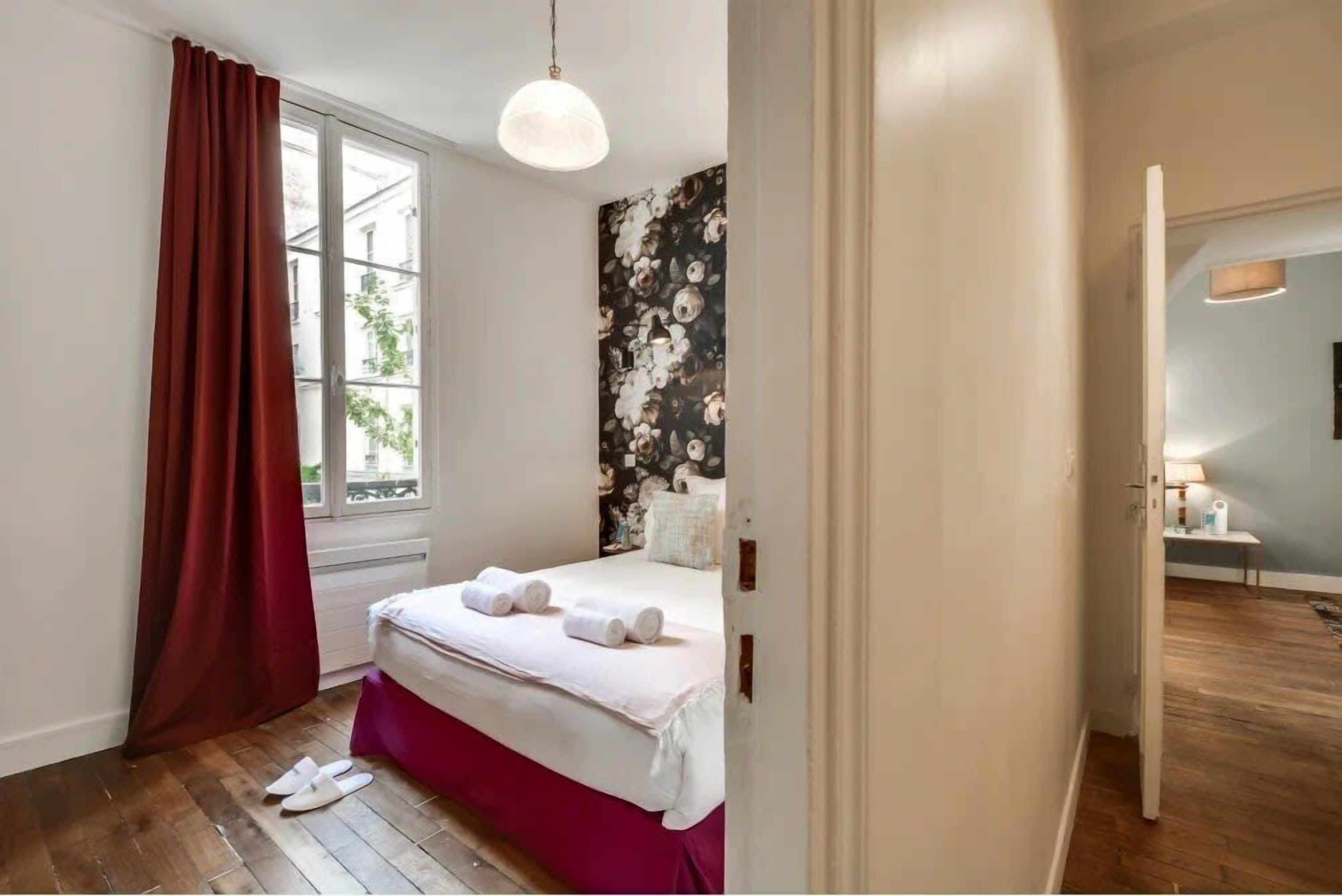 Un dormitorio con cama, una ventana con cortinas, una pared con papel tapiz floral y un pasillo que conduce a otra habitación.